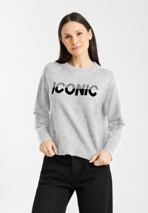 Grijze, fluffy trui met het woord "ICONIC" in zwarte letters. Ronde hals, lange mouwen en een iets verkorte zoom. Gecombineerd met zwarte broek.
