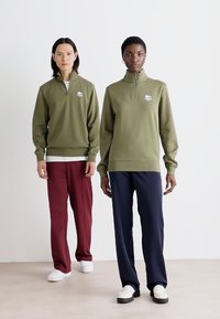 Alie žalios spalvos zip-neck džemperiai su Lacoste logotipu, derinami su bordo ir tamsiai mėlynomis kelnėmis. Džemperiai turi briaunotus rankogalius ir apykaklę.
