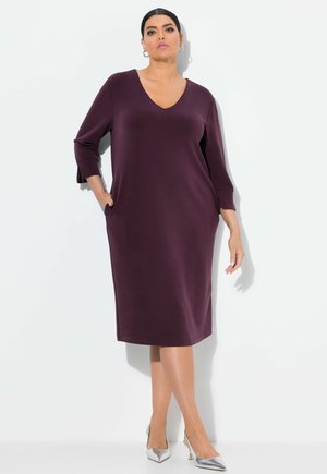 Vestido de manga tres cuartos con escote en V en tela de color púrpura profundo, con un ajuste relajado y bolsillos laterales. Combinado con zapatos de tacón alto plateados.