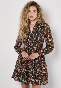 Langärmliges, blumiges Kleid in Dunkelgrün. Mit gesmoktem Taillenbereich, rüschensäumigem Saum und einem V-Ausschnitt, verziert mit einer lebhaften Mischung aus orangefarbenen und weißen Blumen.