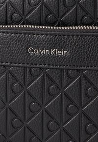 Zwarte leren handtas met een geborduurd geometrisch patroon, een zilveren rits en het Calvin Klein-logo in een moderne lettertype.