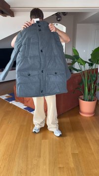 Persona che tiene un cappotto imbottito grigio con bottoni e tasche in un soggiorno con pavimento in legno e una grande pianta in vaso.