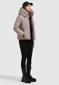 Gai bēšas krāsas pūkainais jakas ar augstu apkakli, sānu kabatām un priekšējo rāvējslēdzēju, saskaņota ar melnajām zeķubiksēm un ceļa augstuma šņorējamām zābaciņām.