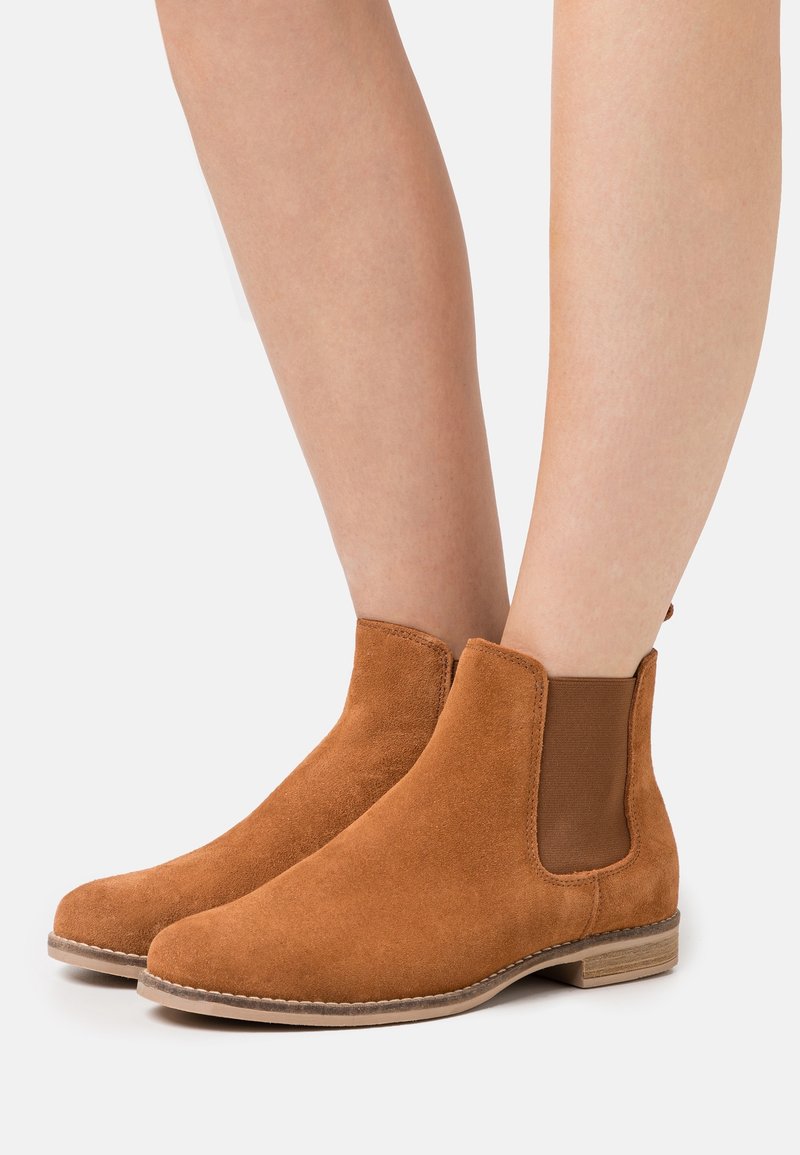Bottines en suède marron avec panneaux latéraux élastiques portées sur jambes nues sur fond clair uni.