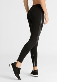 Osoba oblečená v černých legínách s bočním reflexním pruhem a černých teniskách s bílou podešví, stojící na jednobarevném pozadí.