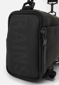 Borsa rettangolare nera realizzata in materiale opaco, caratterizzata da logo "RAINS" in rilievo, chiusura con zip e tracolla regolabile con clip. Superficie testurizzata.