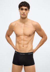 Černé bavlněné boxerské trenky s elastickým pasem, na kterém je bílé logo "Calvin Klein", nošeny modelem s atletickou postavou.