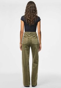 Sieviete ar gariem viļņainiem matiem, ģērbusies melnā pieguļošā īsām piedurknēm blūzē un augsto vidukli akmens zaļās platajās biksēs, stāv ar seju prom no skatītāja.