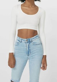 Frau in einem weißen langärmeligen, kurzen Rippoberteil und hoch taillierten, hellblauen, ausgefransten Jeans, die vor einem schlichten Hintergrund steht.