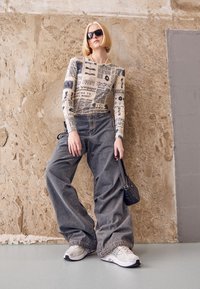 Långärmad beige topp med tidningstryck, kombinerad med överdimensionerade grå denimjeans och vita sportiga sneakers. Svart handväska kompletterar stilen.