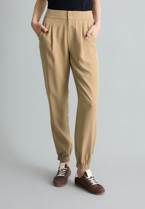 Vrouw draagt beige broek met elastische taille en taps toelopende pijpen met elastische boorden, bruine veter sneakers, handen in zakken, staand op een grijze vloer.