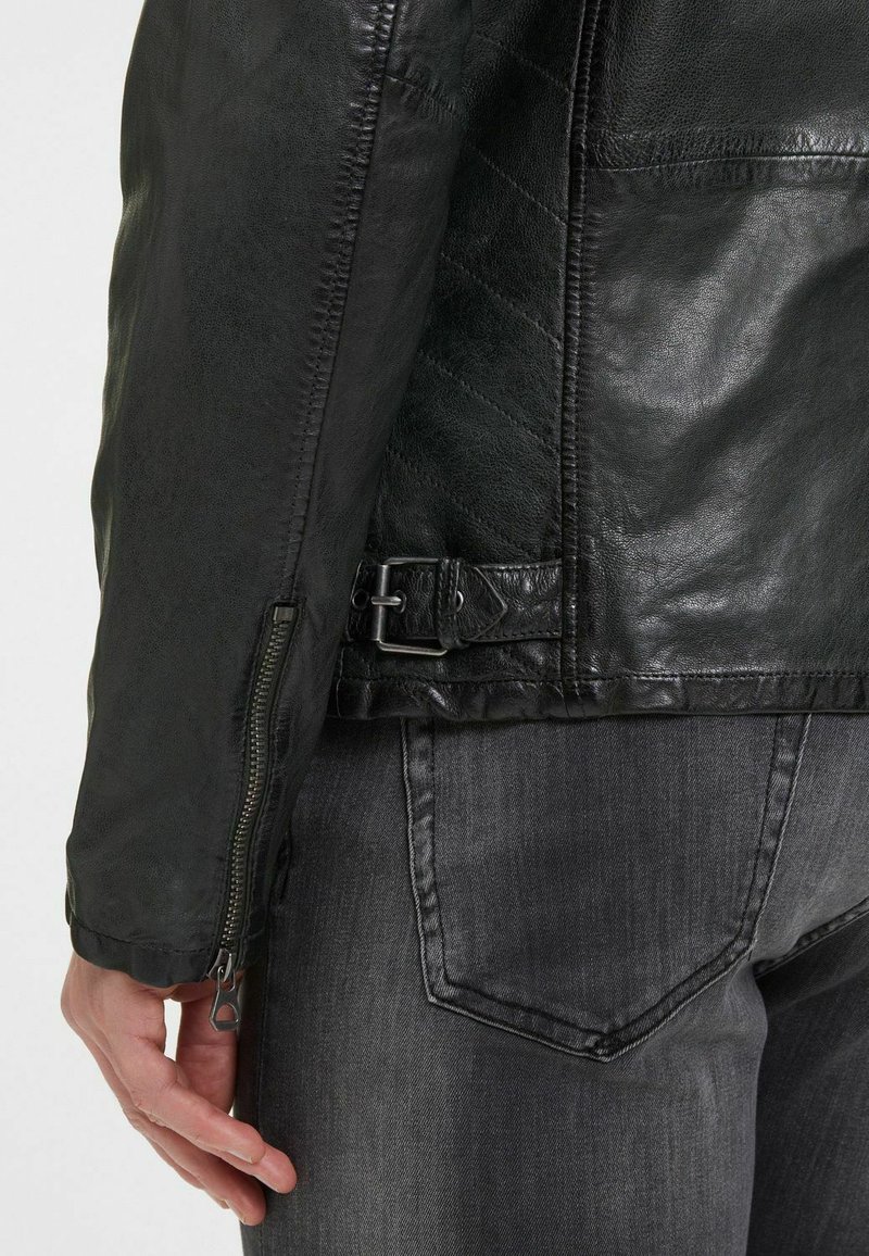 Chaqueta de motociclista de cuero negro con puño con cremallera, hebilla lateral ajustable y acabado texturizado, combinada con jeans de mezclilla grises.