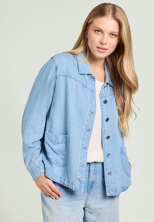 Młoda kobieta o długich blond włosach, ubrana w jasnoniebieską kurtkę dżinsową na białym topie oraz jasnoniebieskie dżinsy ozdobione małymi cyrkoniami.