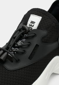 Black mesh sneaker with black elastic laces and toggle, white sole, and a white label with partially visible black text on the tongue.