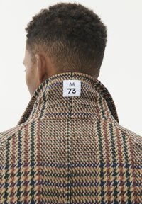 Personas skats no aizmugures, tērpts augsta apkakles brūnā šallē ar rūtainu rakstu, uz apkakles ir balta etiķete ar uzrakstu "M 73".