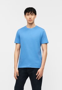 Νέος άνδρας με κοντά μαύρα μαλλιά, φορώντας μια απλή ανοιχτό μπλε κοντομάνικη μπλούζα και σκούρο τζιν, στέκεται μπροστά σε ένα λευκό φόντο.