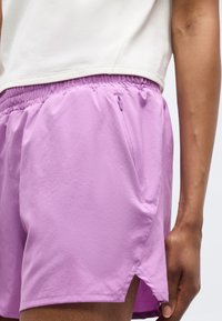 Κοντινό πλάνο ατόμου που φοράει λιλά σορτς με ελαστική μέση, πλαϊνή τσέπη και μικρή σκίσιμο στο τελείωμα, σε συνδυασμό με ένα κοντό λευκό τοπ.