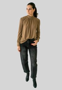 Vrouw met donker haar in een paardenstaart, gekleed in een bruine blouse met lange mouwen, zwarte jeans en zwarte enkellaarsjes, staand met één hand in de zak.