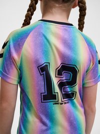 Dziecko z brązowymi, zaplecionymi włosami, ubrane w sportową koszulkę w paski w kolorach tęczy z numerem 12 i napisem "LIGHT UP THE WILD" na plecach.