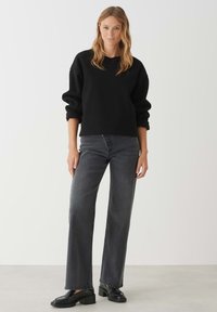 Zwarte sweatshirt met een ronde hals, opgerolde mouwen; gecombineerd met hooggeknoopte grijze jeans en zwarte chunky schoenen op een neutrale achtergrond.