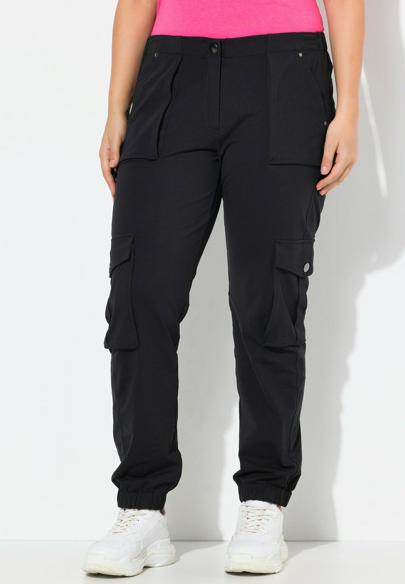 Schwarze Cargo-Hosen aus weichem Material, ausgestattet mit zwei Fronttaschen, gerippten Bündchen und Druckknopf-Details. Getragen mit weißen Sportschuhen.
