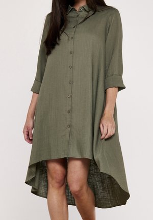 Vrouw die een olijfgroene overhemdjurk draagt met driekwarts mouwen opgerold en een hoog-laag zoom, staande tegen een effen achtergrond.