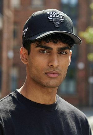 Man with curly hair wearing black Chicago Bulls cap and black shirt, standing outdoors in blurred urban setting.
