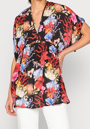 Femme portant un chemisier noir avec imprimé tropical floral coloré et corail, col en V, et manches courtes, associé à un pantalon blanc.
