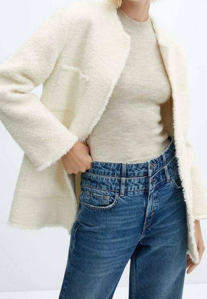 Abrigo de textura crema con el frente abierto; suéter de lana beige debajo; jeans de mezclilla azul de tiro alto con bolsillos frontales y acabado desgastado.