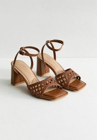 Brown heeled sandals with a square toe, woven strap detail, adjustable ankle strap, and a chunky block heel. Smooth leather finish.