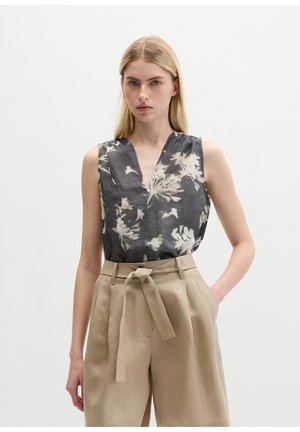 Femme portant un chemisier sans manches gris foncé à motif floral, rentré dans un pantalon beige taille haute avec une ceinture nouée, debout sur un fond uni.