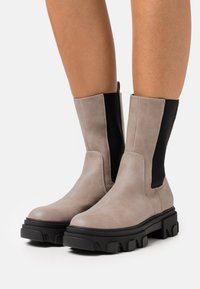 Lila mid-calf stövlar med chunky svarta sulor och elastiska svarta sidopaneler, barfota, mot en enkel vit bakgrund.