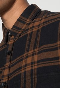 Camicia a quadri nera e marrone, caratterizzata da un colletto, bottoni neri e un tessuto morbido e testurizzato con linee orizzontali e verticali ben definite.