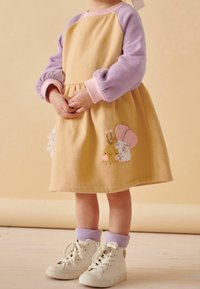 Enfant portant une robe pastel avec des manches violettes et une appliqué escargot, assortie de baskets blanches et de chaussettes violettes, se tenant sur un sol clair.