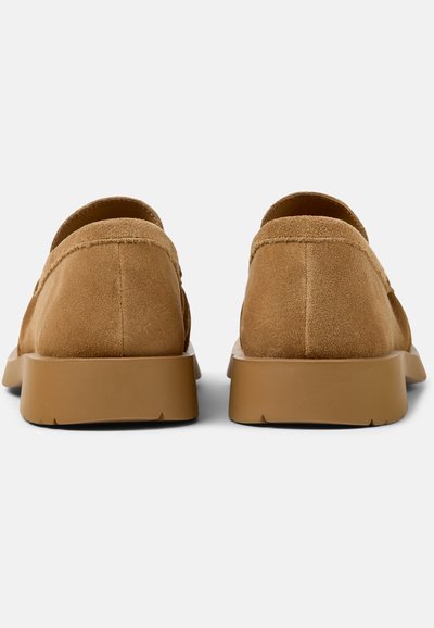 Paar hellbraune Wildleder-Slipper mit glatten Gummisohlen, von hinten auf weißem Hintergrund gezeigt.