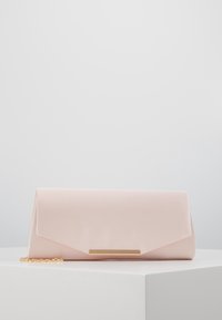 Jasnoróżowa satynowa kopertówka z złotym łańcuszkiem i złotym prostokątnym zapięciem leżąca na białej powierzchni na tle jasnoszarym.