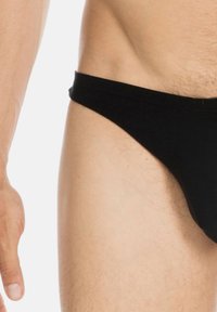 Schwarzer Stoff-Tanga mit glatter Textur, niedrig geschnittenem Design und einem minimalen Bund. Nahaufnahme des unteren Körperbereichs, die Haut und Körperbehaarung zeigt.