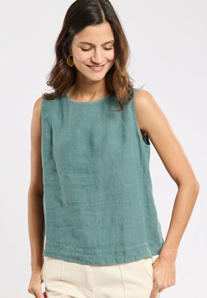 Femme aux cheveux bruns mi-longs, portant des boucles d'oreilles en or, un débardeur bleu sarcelle et un pantalon beige clair, souriante et regardant vers le bas.