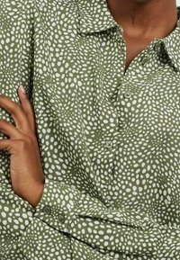 Person som bär en grön skjorta med knappar och ett oregelbundet mönster av vita prickar, med armarna korsade vid midjan.