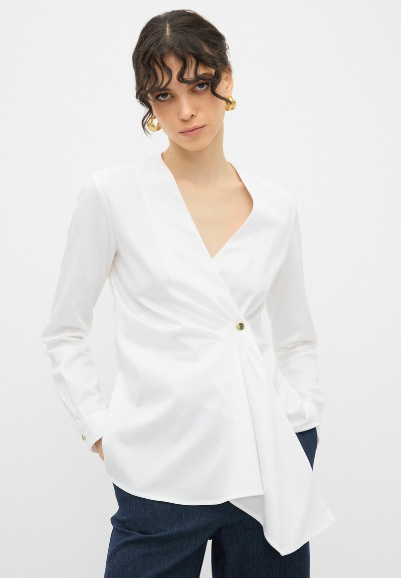 Blusa bianca a maniche lunghe con profondo scollo a V, dettaglio frontale attorcigliato e accento con un singolo bottone. Tessuto morbido e testurizzato; abbinata a jeans scuri.