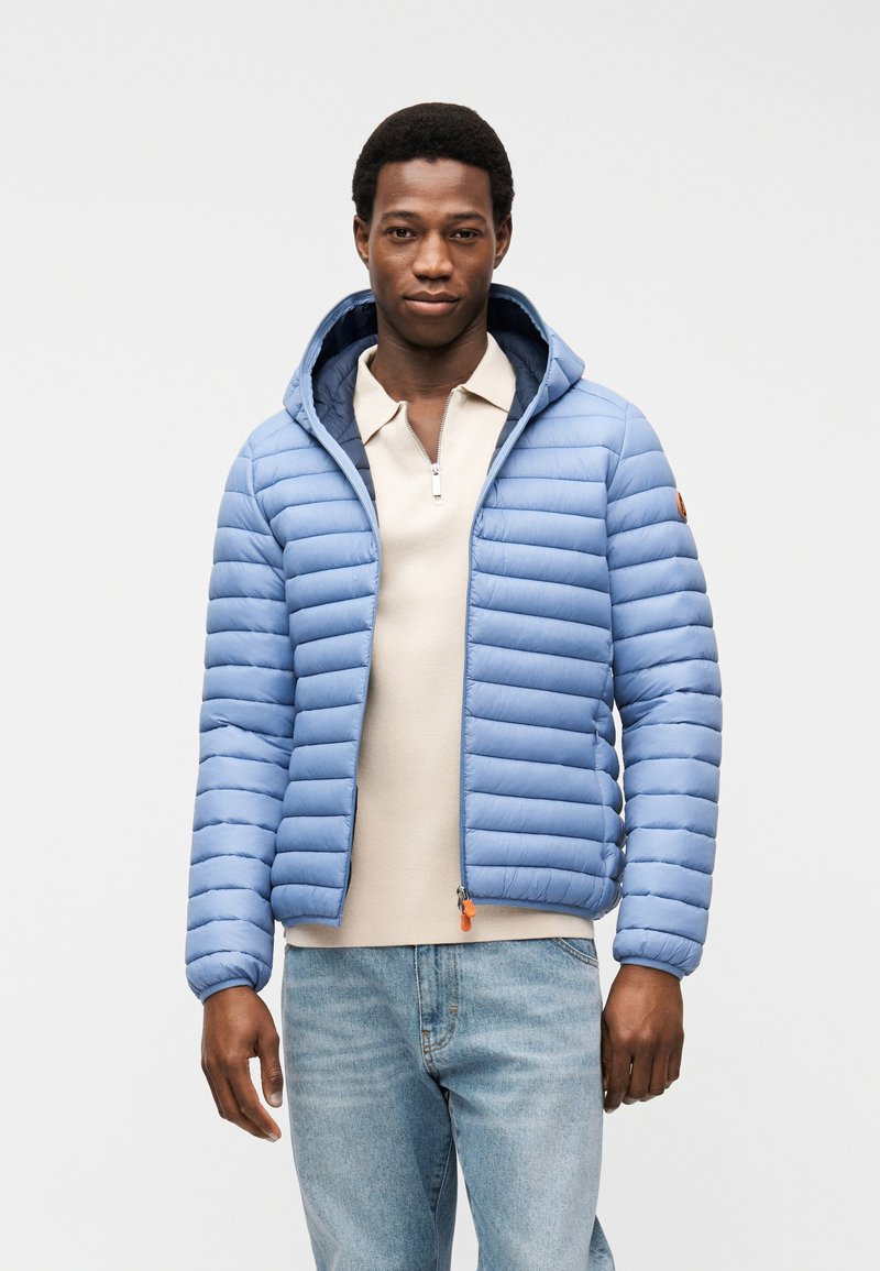 Άνδρας που φοράει ένα γαλάζιο μπουφάν με επένδυση πάνω από μια μπεζ ζακέτα με φερμουάρ και γαλάζια τζιν, στέκεται μπροστά σε απλό λευκό φόντο.