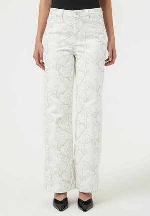 Bijele hlače širokih nogavica s diskretnim svijetlosivim uzorkom cvjetnih vinove loze, nošene s crnim šiljastim cipelama i crnim topom.