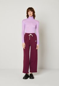 Gaiši violetas turtlenecks ar garām piedurknēm; bordo platās bikses ar aukliņu jostasvietā; melni slip-on apavi; minimālistisks fons.