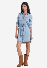 Ljusblå denimskjortklänning med långa ärmar, två bröstfickor, midjebälte och rundad fåll, kombinerad med bruna cowboyboots.