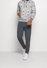 Grauer Hoodie mit einem sich wiederholenden schwarzen Logomuster, kombiniert mit dunkelgrauen, schmal zulaufenden Jogginghosen mit einem Logo-Patch und elastischen Bündchen, getragen mit weißen Sneakers.