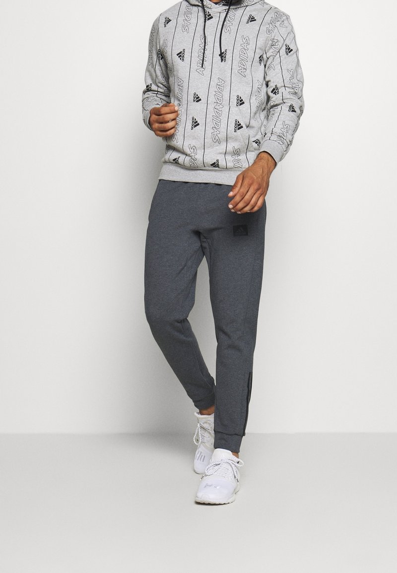 Grauer Hoodie mit einem sich wiederholenden schwarzen Logomuster, kombiniert mit dunkelgrauen, schmal zulaufenden Jogginghosen mit einem Logo-Patch und elastischen Bündchen, getragen mit weißen Sneakers.