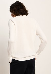 Person mit kurzem lockigem Haar, der einen weißen, langärmeligen, strukturierten Pullover und schwarze Hosen trägt und mit den Händen in den Taschen von hinten steht.