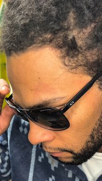 Juodos akiniai nuo saulės su blizgiu paviršiumi, ant šoninės dalies matomas išskirtinis logotipas (A/X), laikomi arti veido priešais tekstūruoto audinio foną.