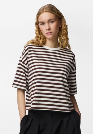 Ung kvinde med krøllet blondt hår iført en brun og hvidstribet t-shirt og sorte bukser, hænderne i lommerne, neutral udtryk.