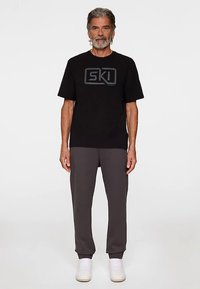 Musta puuvillainen t-paita, jossa on harmaa "SKI"-logo, yhdistetty harmaisiin verkkareihin ja valkoisiin lenkkareihin. Malli seisoo suorassa yksivärisen taustan edessä.